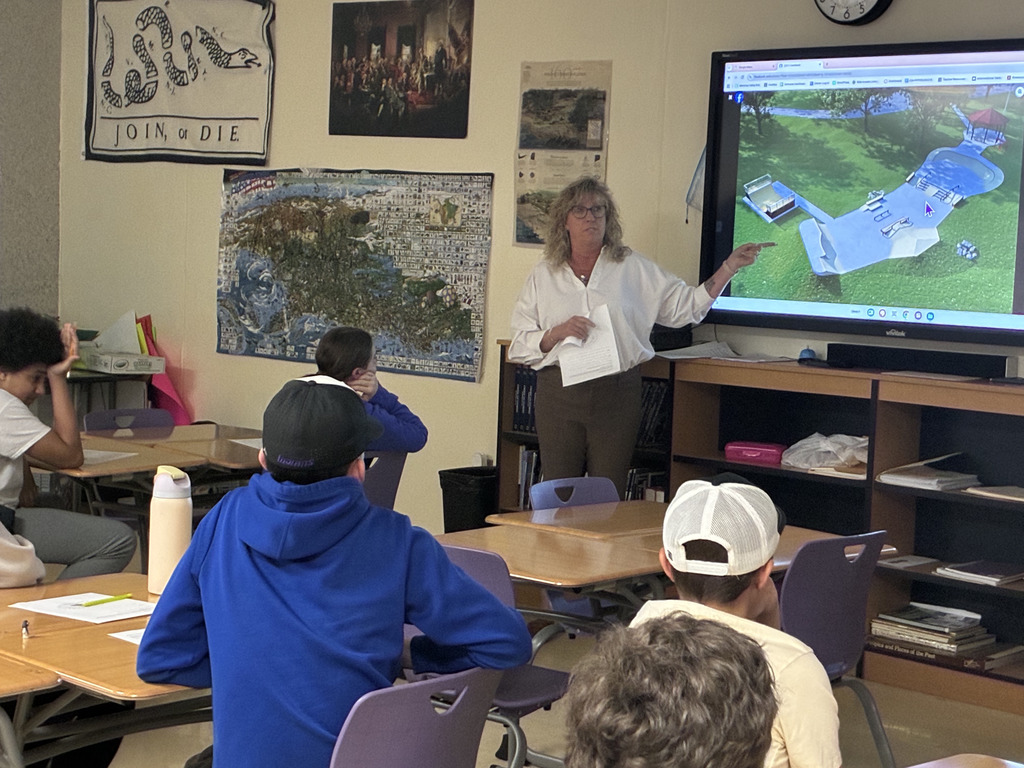 Adult in a white button up standing before a classroom of students giving a presentation.
