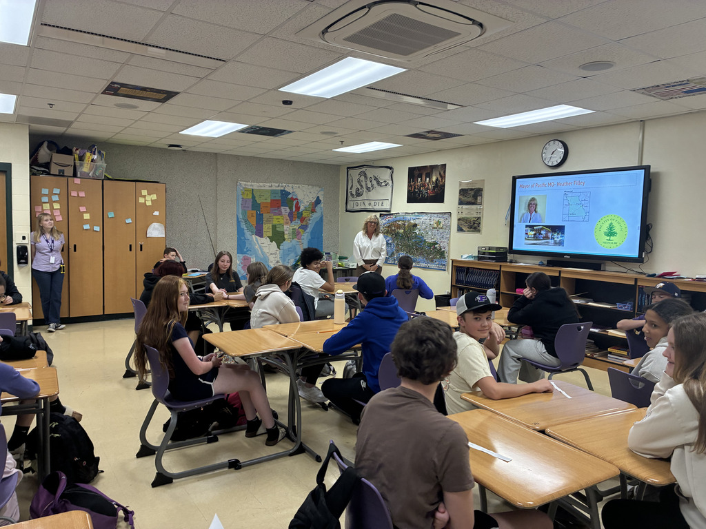 Adult in a white button up standing before a classroom of students giving a presentation.