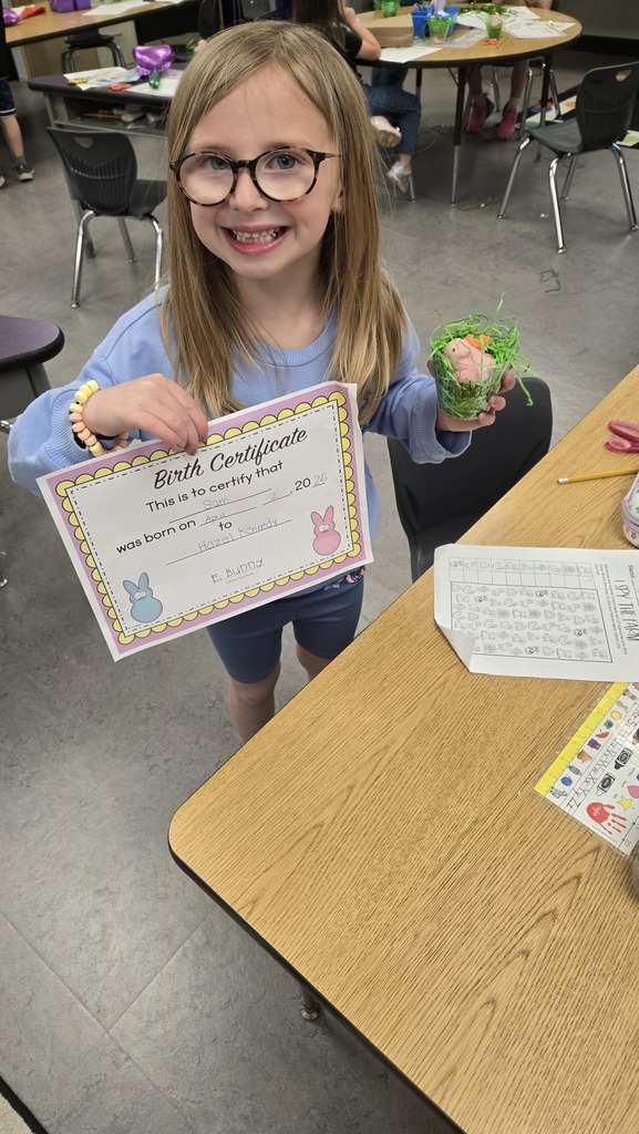 Young student showing a colorful piece of paper and a cup filled with green paper at a classroom desk.