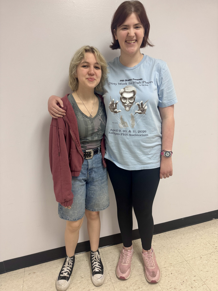 Two students, with one arm around the other, posing for a photo in a hallway.