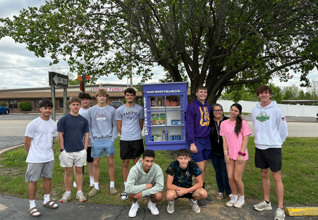Group of students posing in front of a small purple food pantry outdoors.