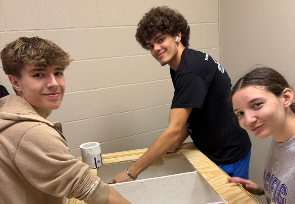 Three students posing for a photo while painting on wood.