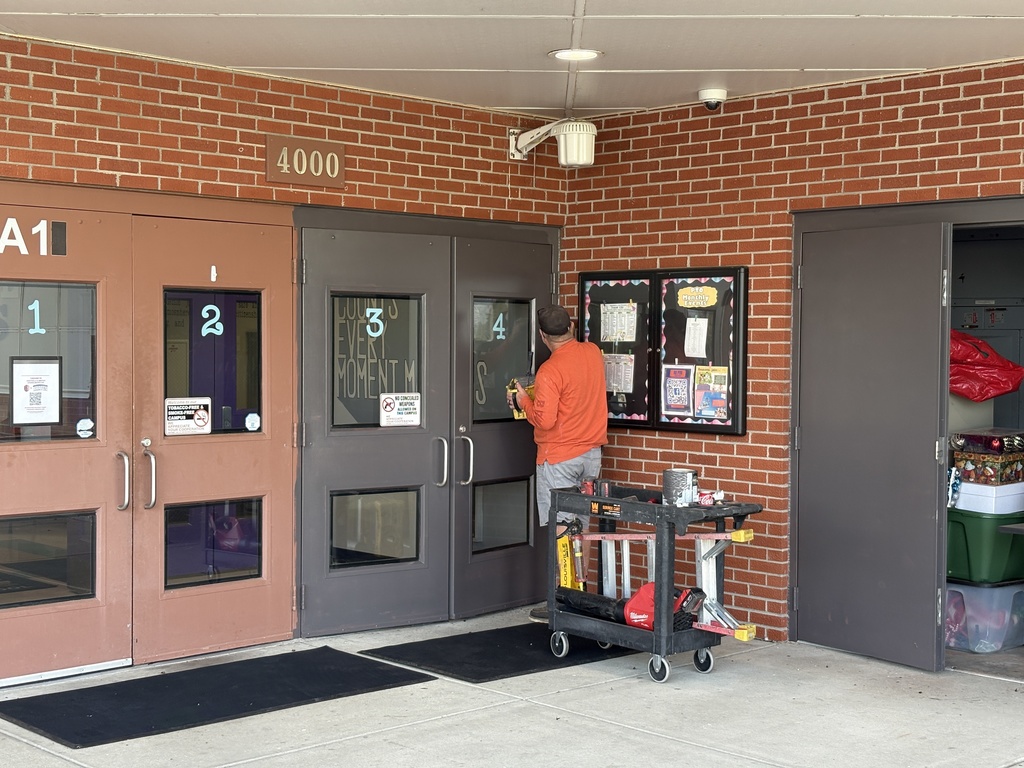 Staff member in orange shirt works on double doors at a brick building entrance, using tools beside a rolling cart.