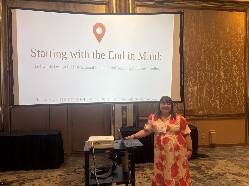 Presenter stands beside a screen displaying “Starting with the End in Mind,” with laptop and projector in a conference room.