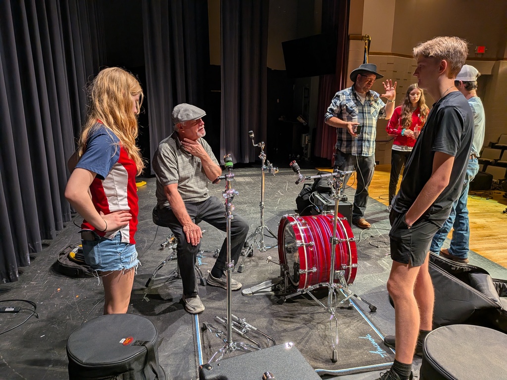 Students talking to a band member on stage with a red drum set in the background.