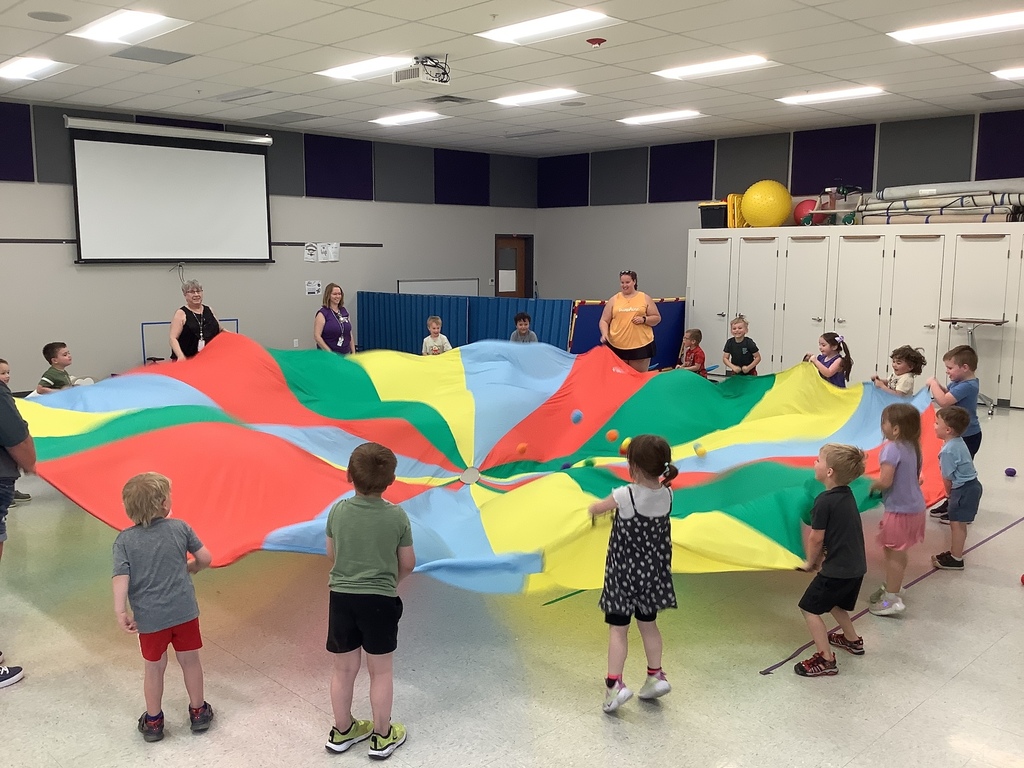 Students and staff hold a colorful parachute while standing around in a circle indoors.