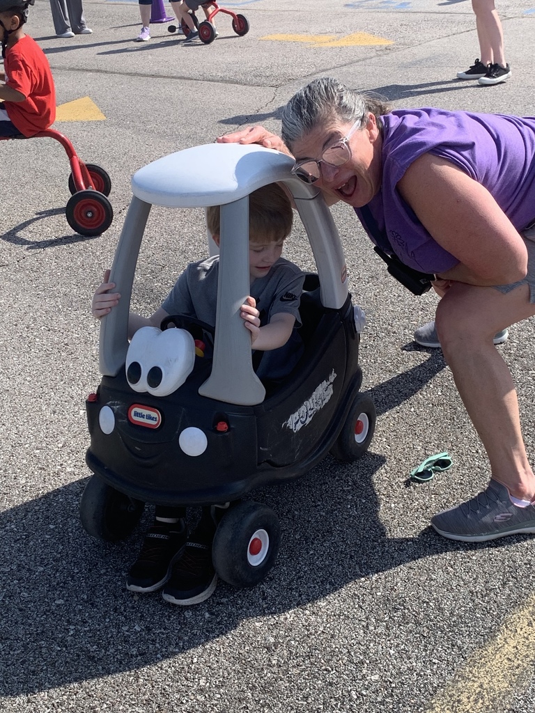 A student drives a toy car, with a teacher nearby smiling.