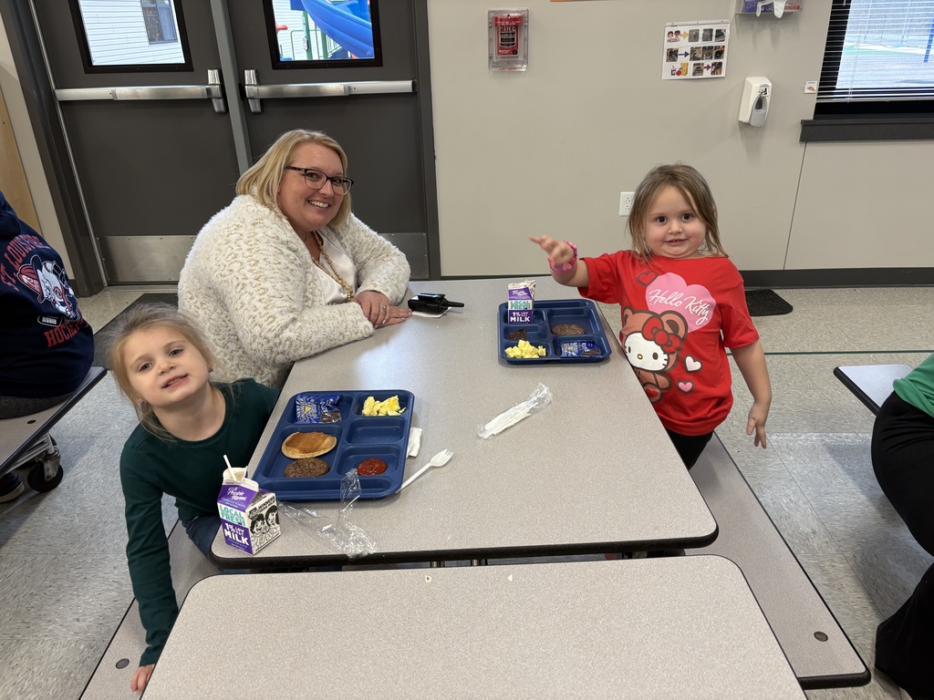 Adult and two young girls sit at a cafeteria table with school lunches; one child smiles while another points, wearing a red Hello Kitty shirt.
