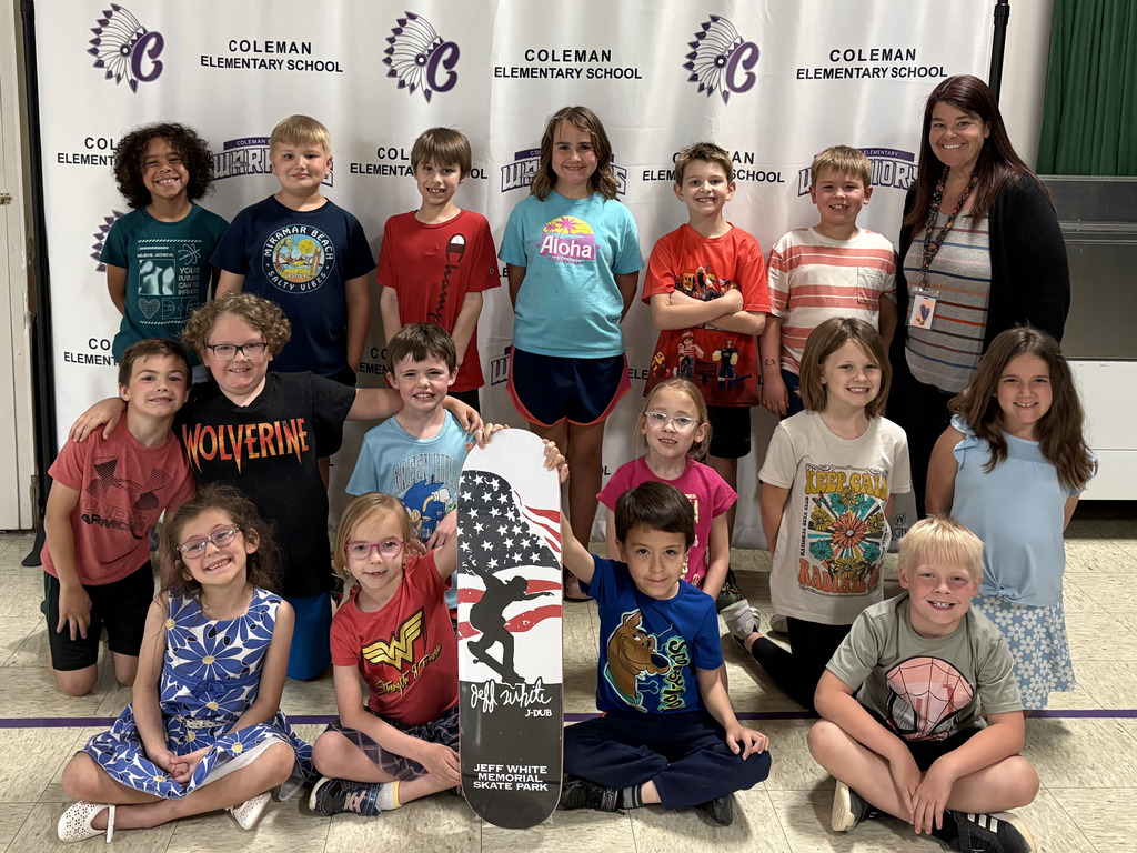 A group of students and staff member stand pose for photo in front of a large white banner while holding a skateboard.