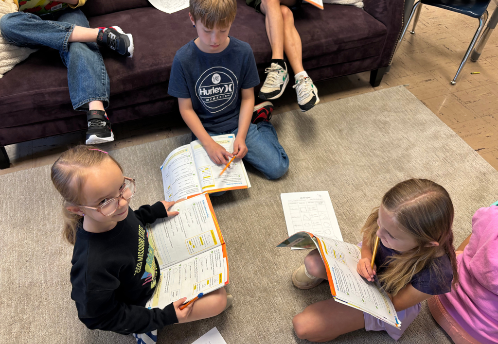Three students sitting on a classroom floor reading school packets.