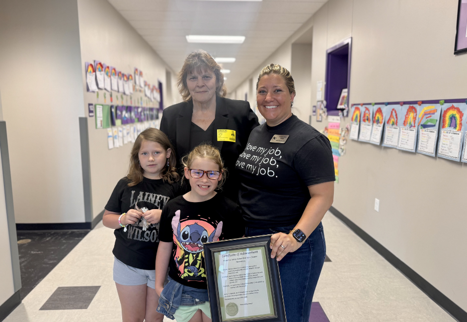 Two students and two adults holding a paper certificate in a black picture frame.