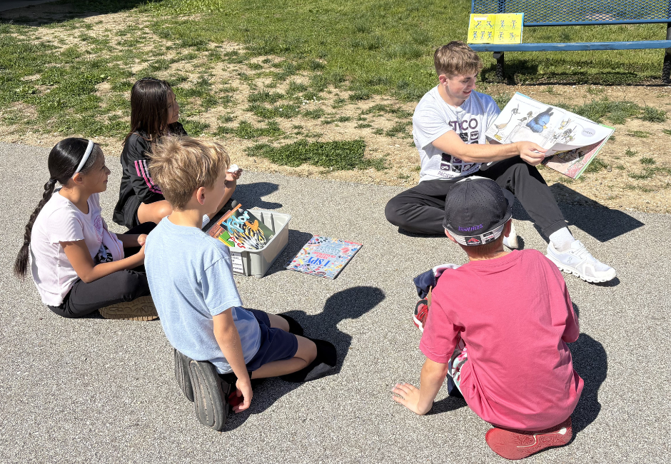 High school student reads to a group of students outside.
