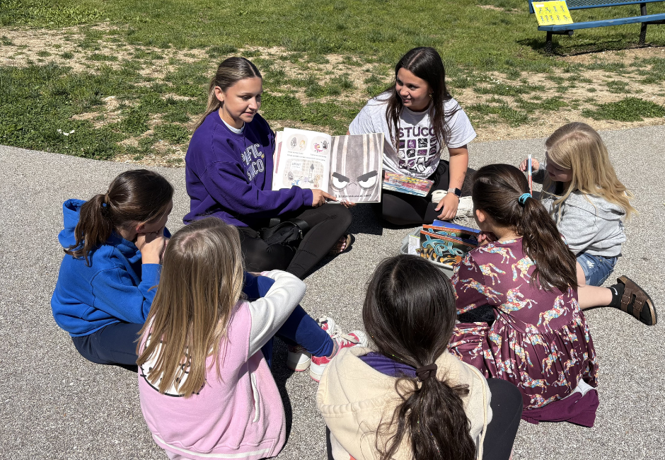 Two staff members read to a group of students outside.