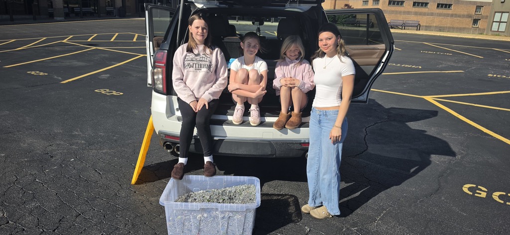 Four girls sit on the open trunk of an SUV in a sunny parking lot, with a clear bin of collected can tabs in front.