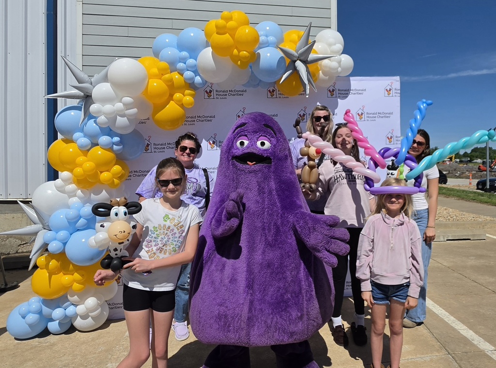 Group poses with purple mascot Grimace and children in balloon hats beneath a colorful arch at a Ronald McDonald House event.
