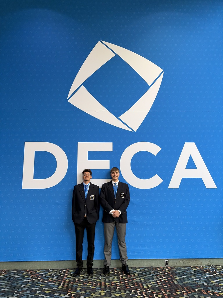 Two students wearing suits stand in front of a large blue "DECA" wall.