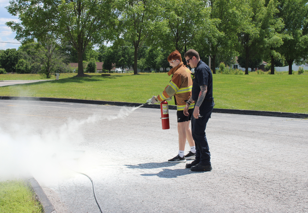 Student sprays a fire extinguisher while a fireman assists. 
