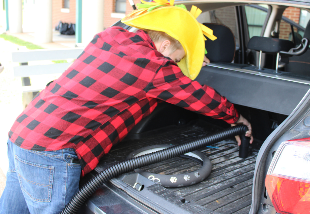 Student wearing a large yellow hat using a vacuum to clean a car trunk.