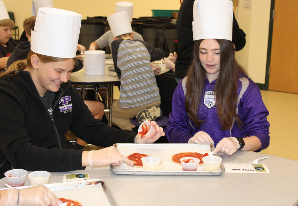 Two students wearing chef hats make pizza in a school cafeteria.
