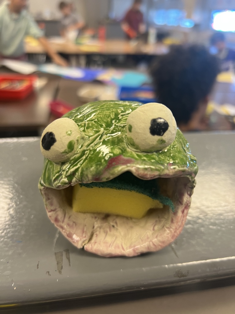 Green ceramic frog-shaped sponge holder with wide eyes and a sponge tucked inside its open mouth in a classroom setting.