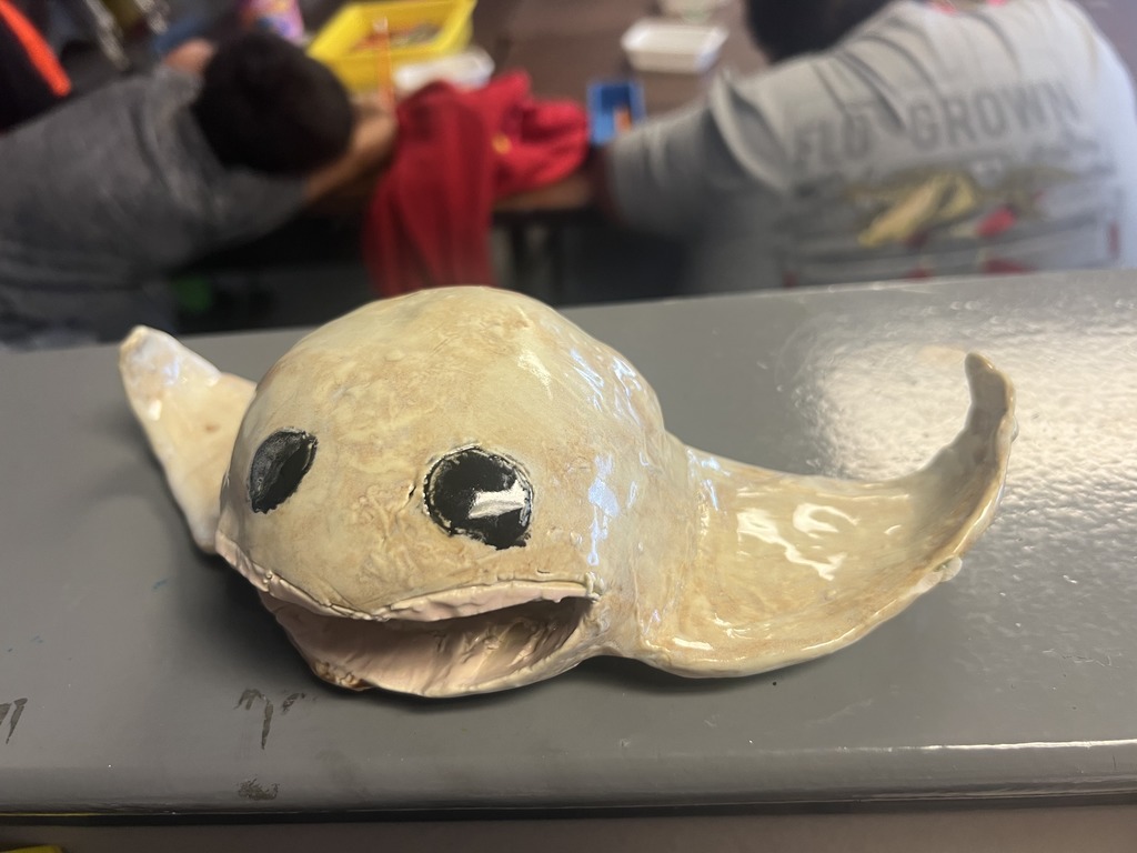 White ceramic sea creature sculpture with black eyes, open mouth, and curved fins displayed on a classroom table.