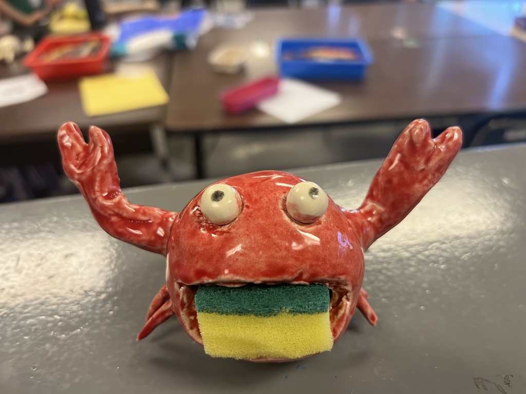Red ceramic crab-shaped sponge holder with raised claws and a yellow-and-green sponge held in its mouth.