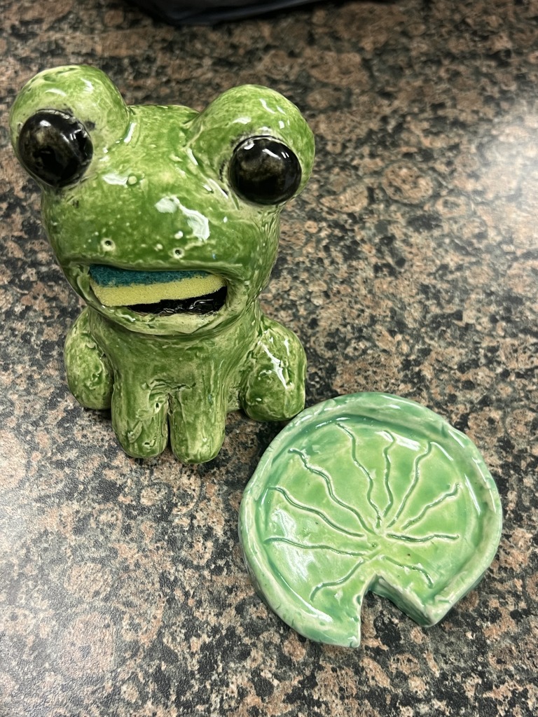 Green ceramic frog sponge holder beside a matching lily pad dish on a speckled countertop.
