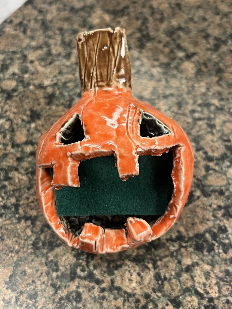 Orange ceramic jack-o’-lantern sponge holder with carved facial features and a green sponge tucked inside its mouth.
