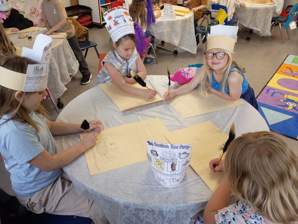 Students wear handmade Boston Tea Party hats while writing with feather pens at classroom tables.
