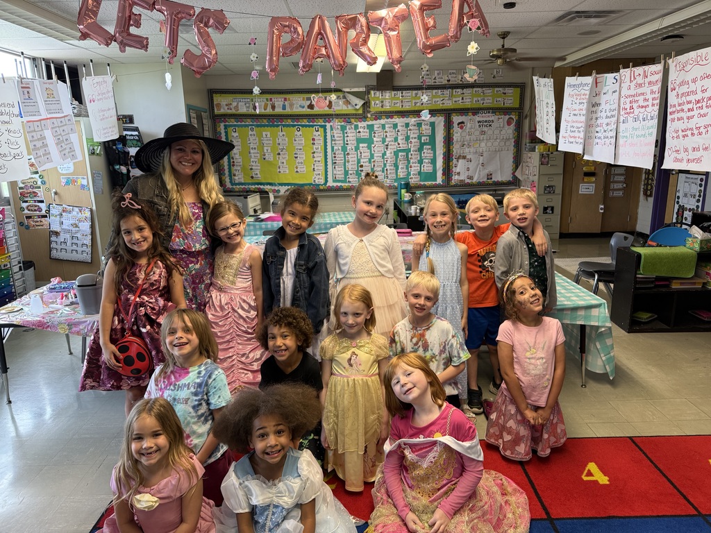Teacher and students dressed in colorful outfits pose together beneath “Let’s Partea” balloons in a classroom.