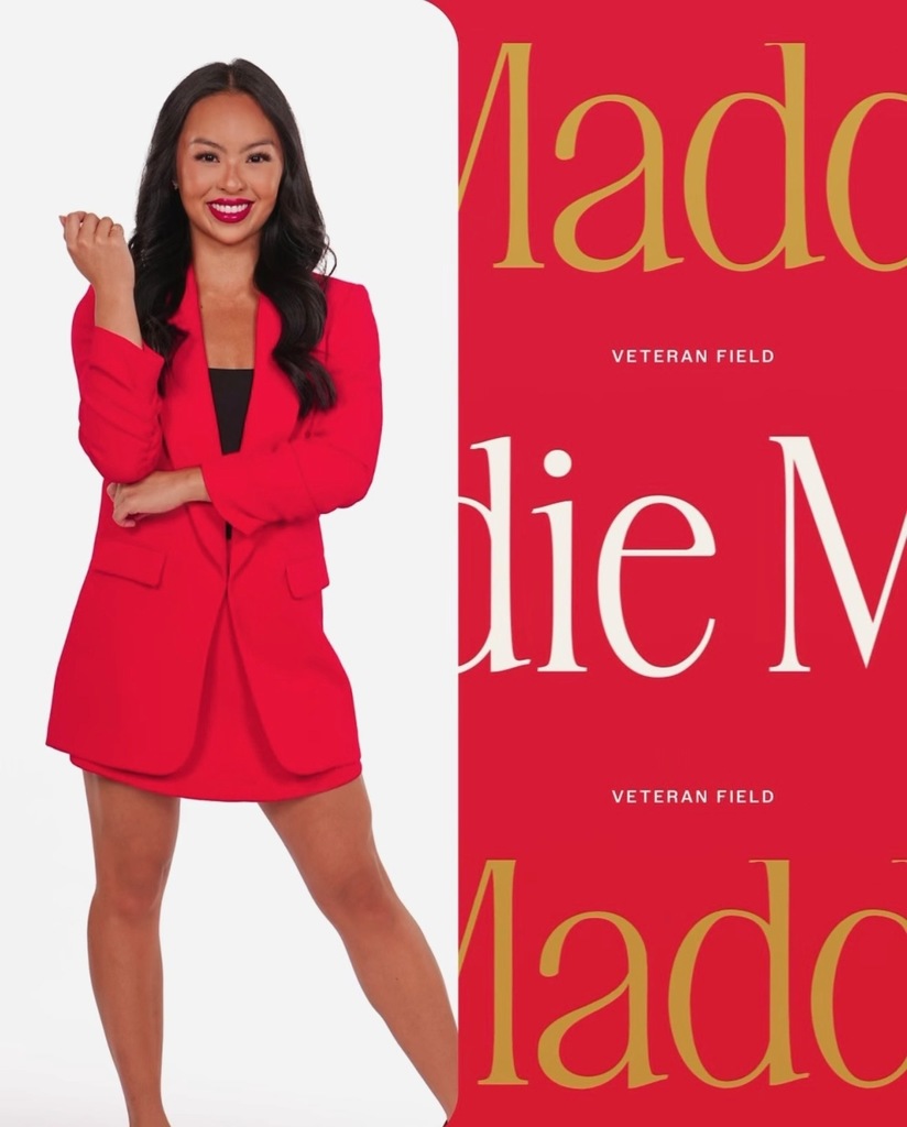 Woman in bright pink blazer dress smiles beside a red backdrop with partial gold lettering.