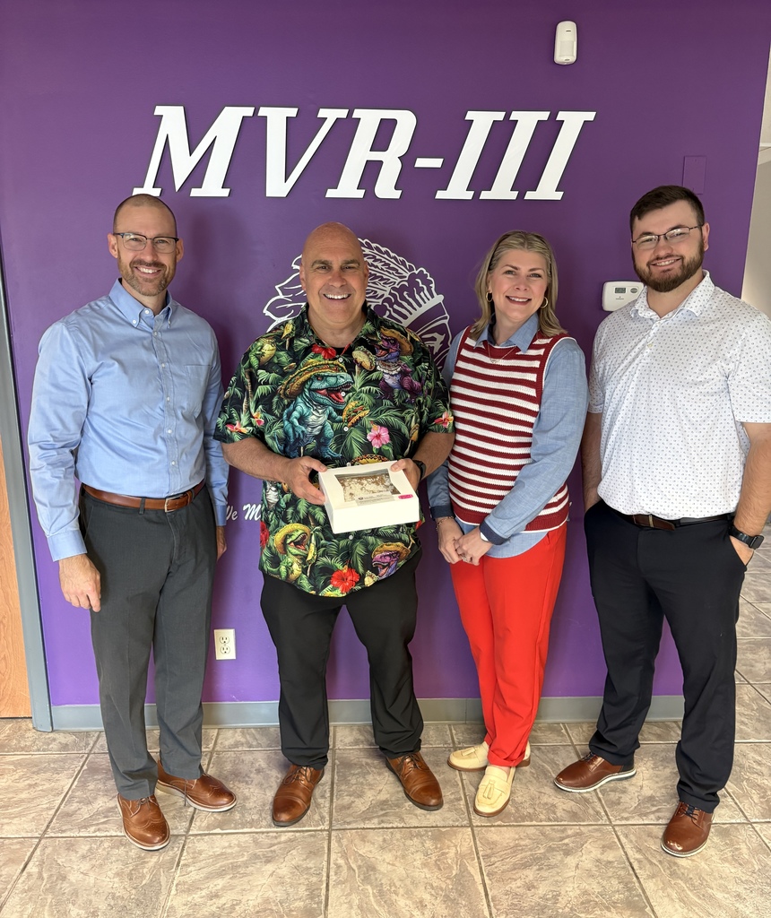 Four adults stand in front of a purple MVR-III wall, smiling as one man holds a boxed dessert.