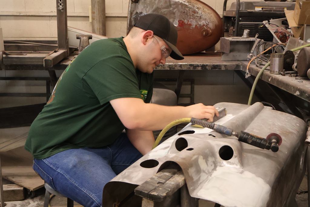Student wearing safety glasses sands a metal vehicle panel in a workshop filled with tools and equipment.