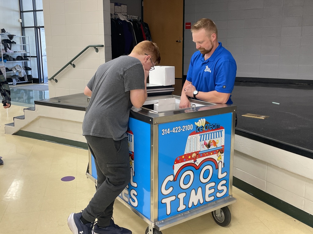 Student leans on a “Cool Times” ice cream cart while speaking with a staff member in a school commons area.