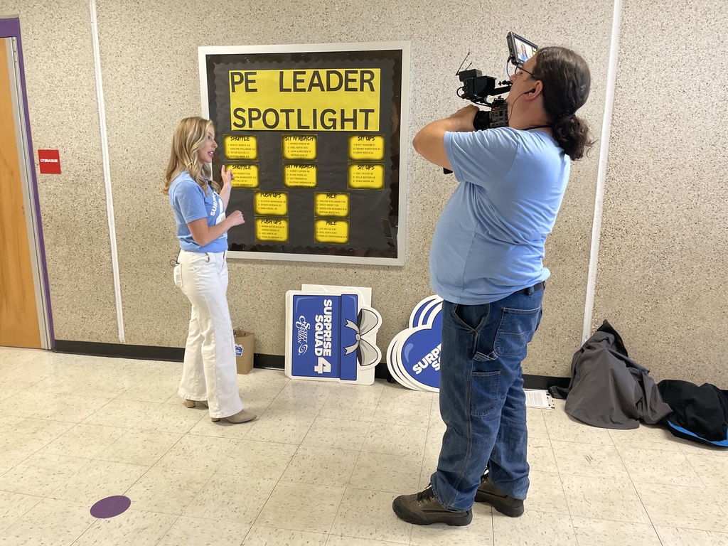 Woman presents a “PE Leader Spotlight” bulletin board while a cameraman films her in a school hallway.