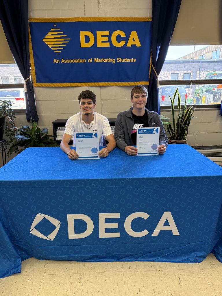 Two DECA students sit behind a blue DECA table holding certificates beneath a DECA banner in a classroom.