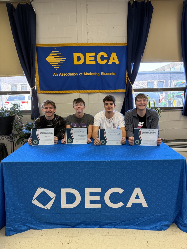 Four DECA students pose with marketing management certificates behind a DECA table and banner display.