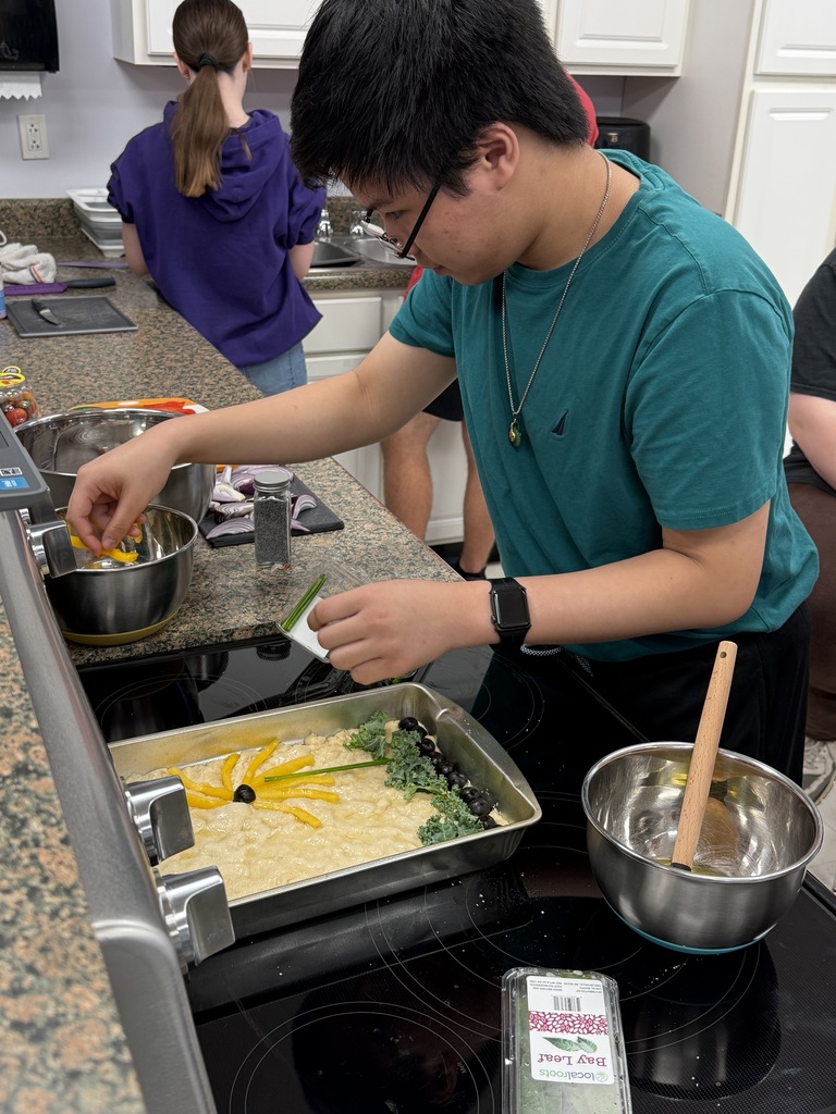 Student decorates focaccia dough with vegetables in a classroom kitchen while classmates prepare food in the background.