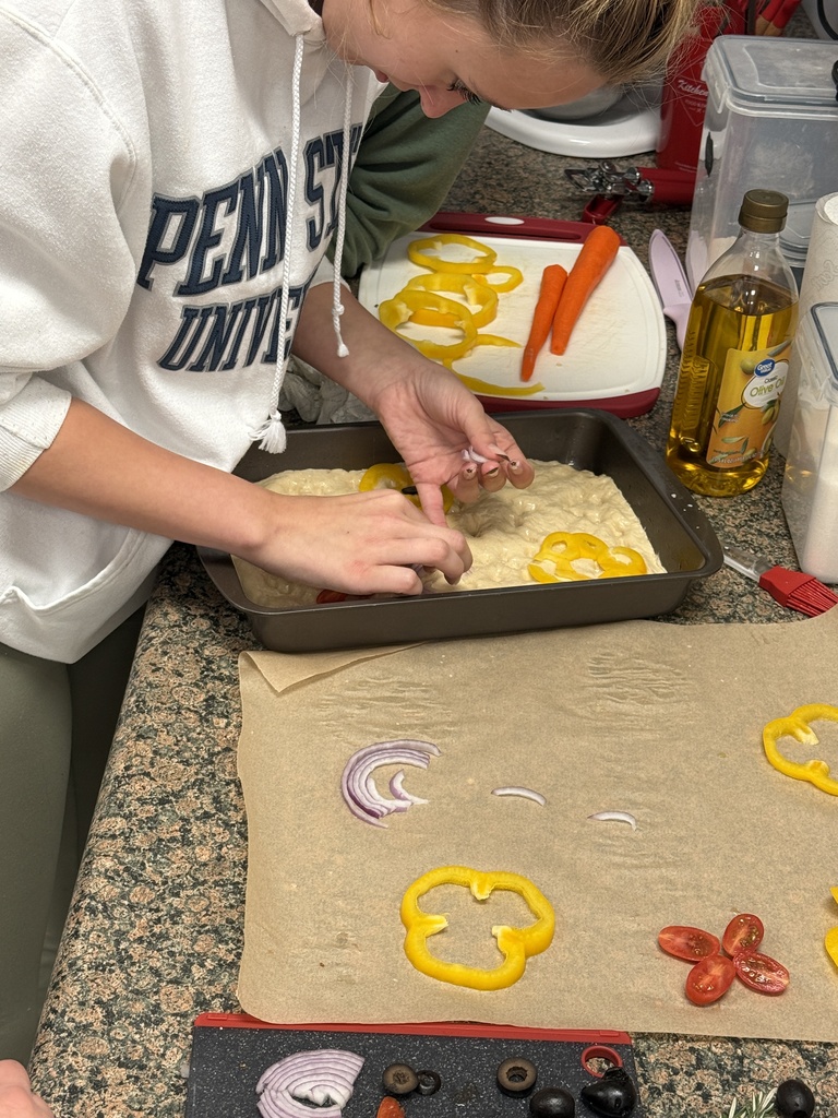 Student arranges sliced peppers, onions, and tomatoes on focaccia dough beside cooking ingredients and utensils.
