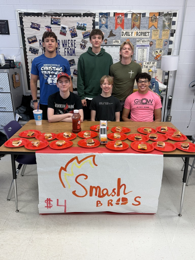 Group of students pose behind a table of mini burgers labeled “Smash Bros” during a classroom food sale.