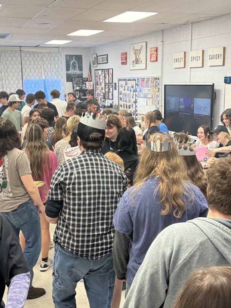 Crowded classroom gathering with students wearing paper crowns and talking during a themed school activity.