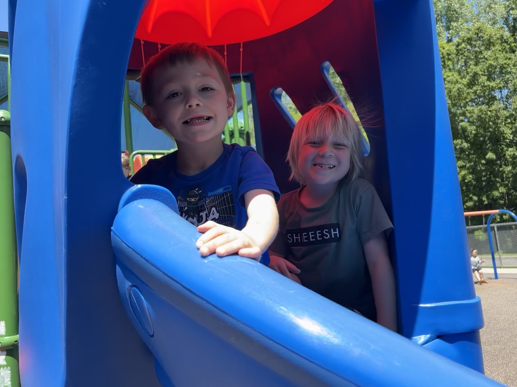 Two students smiling in a blue slide outdoors.