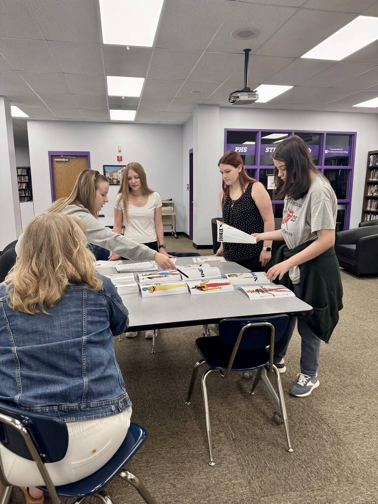 High school students gather around a table in the library, arranging and discussing Egyptian history project materials.