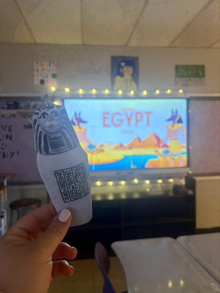 Student holds a handmade Egyptian sarcophagus craft in front of a classroom screen displaying an Ancient Egypt lesson.
