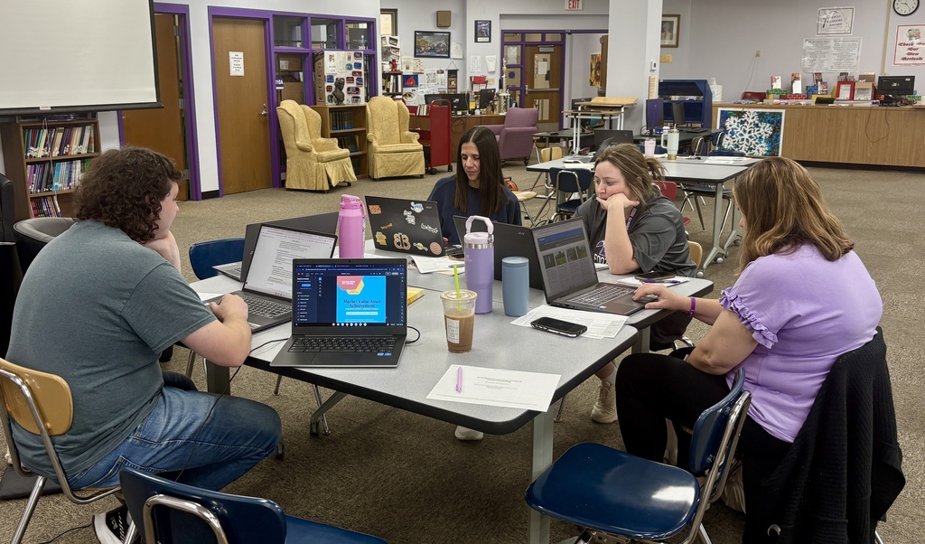 Staff members sit around tables in a school library, collaborating on laptops and reviewing materials during a professional development session.