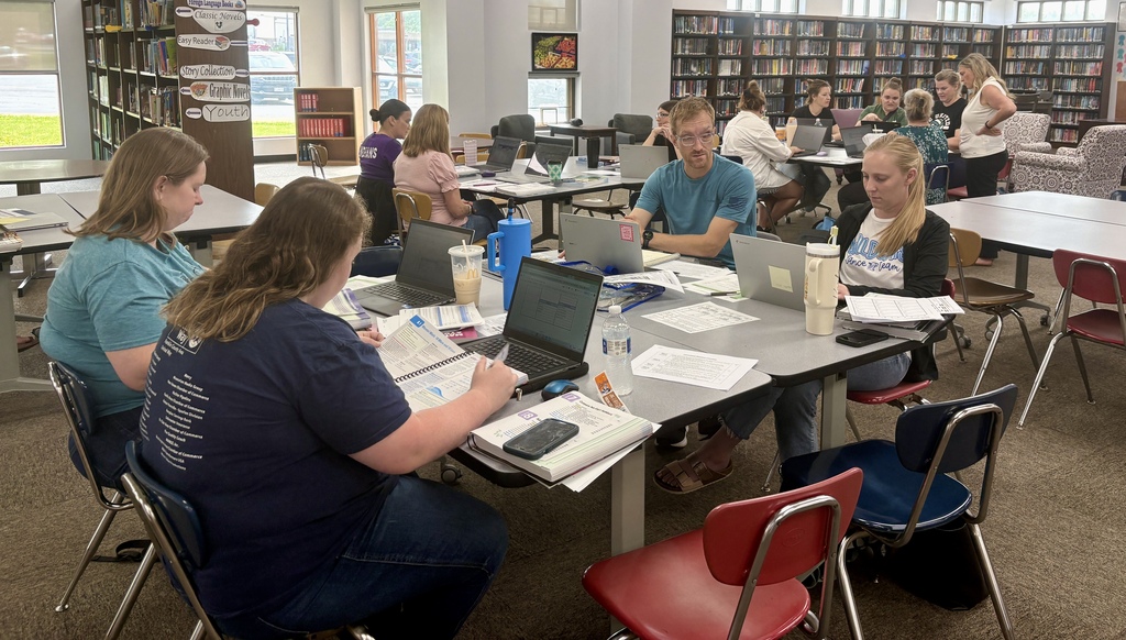 Teachers work together in small groups at tables in a library, using laptops, notebooks, and curriculum materials during training.