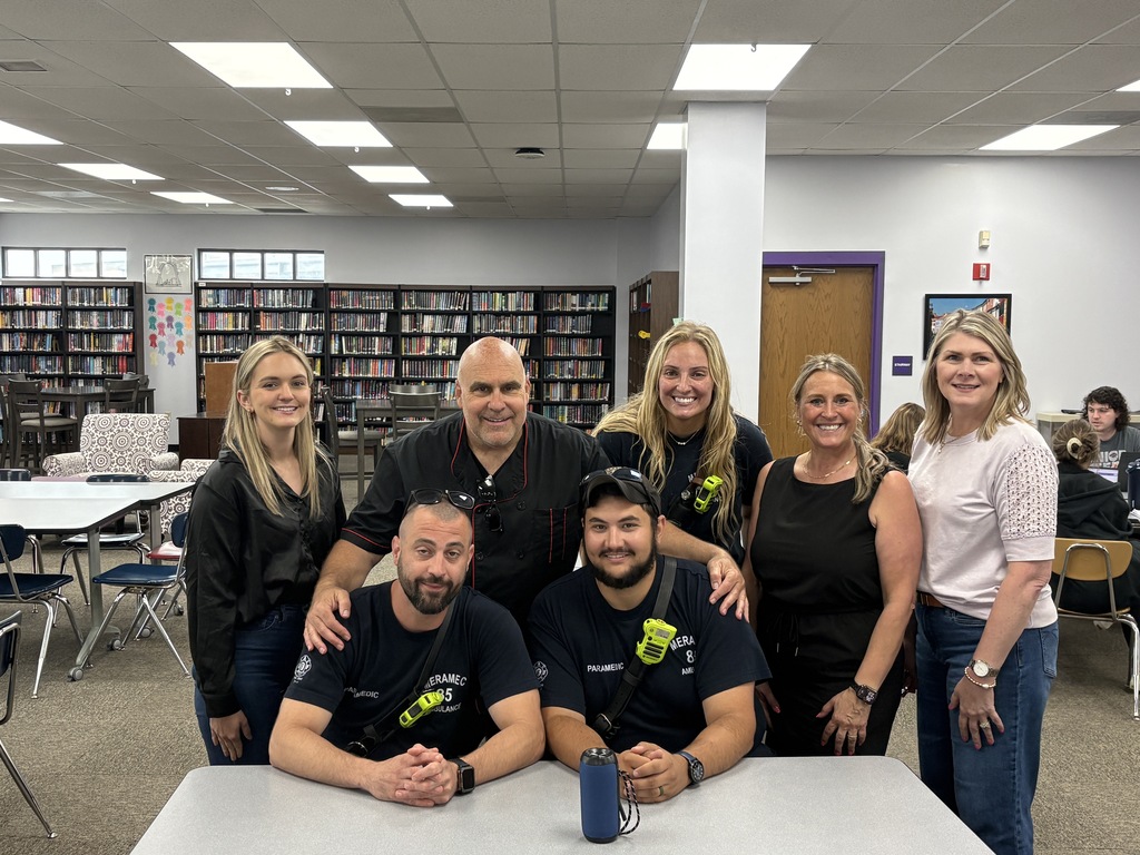 Seven adults, including paramedics, school staff, pose together and smile in a school library with bookshelves and tables in the background.