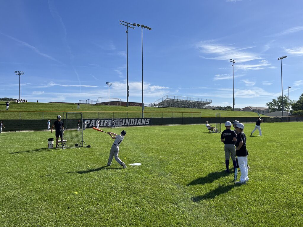 Young baseball player swings at a ball during batting practice while teammates watch from the outfield.