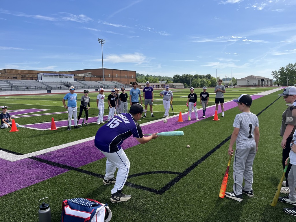 Coach demonstrates a batting drill on a football field as young baseball players watch and wait their turn.