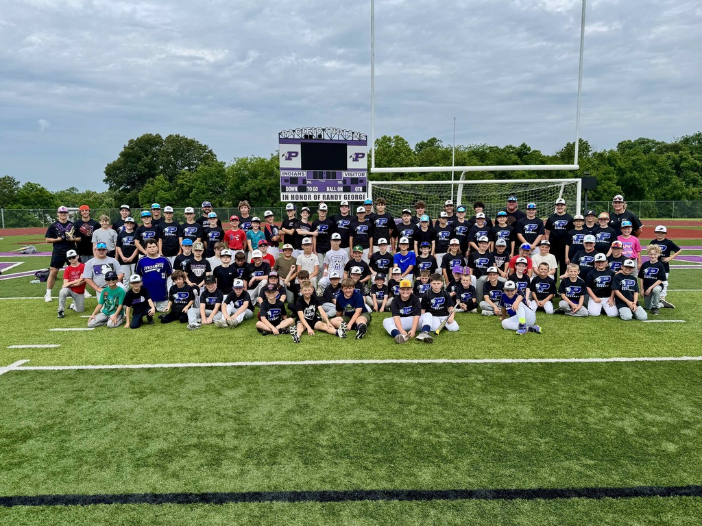 Large group of youth baseball players and coaches pose for a team photo on a football field beneath the stadium scoreboard.